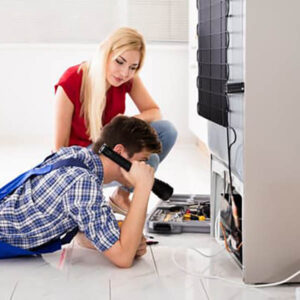 Woman Looking At Male Worker Repairing Refrigerator In Kitchen Room